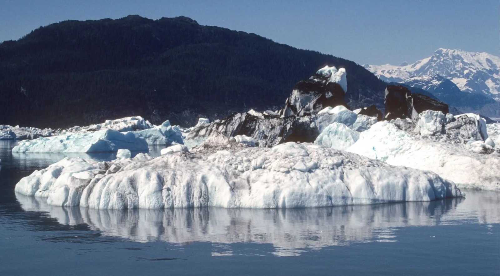 A large grounded iceberg reflected in still water beneath a dark mountain