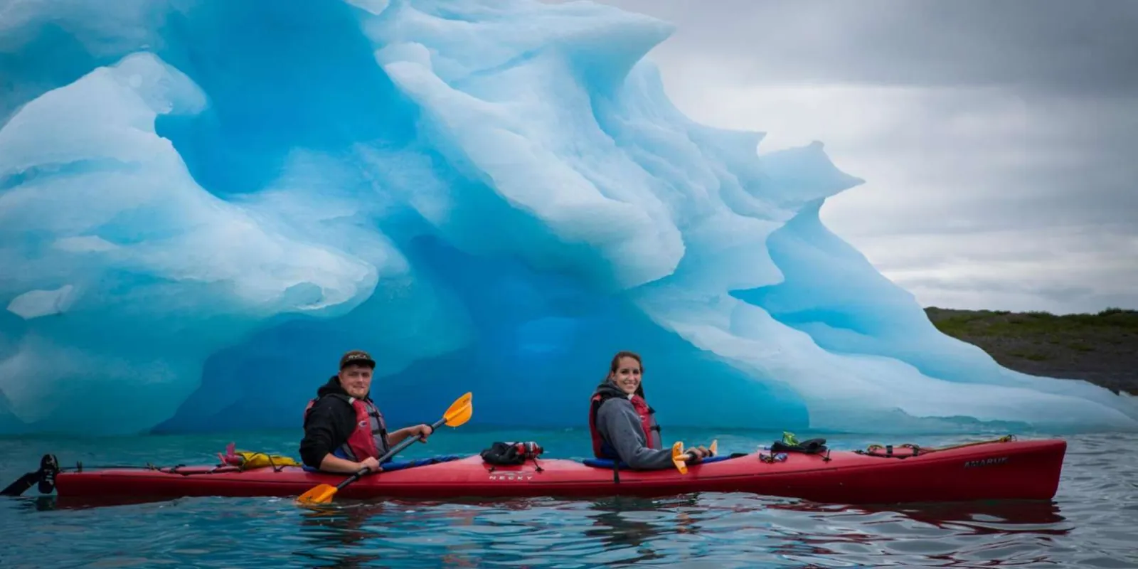 Two kayakers alongside a deep-blue iceberg sculpted by meltwater