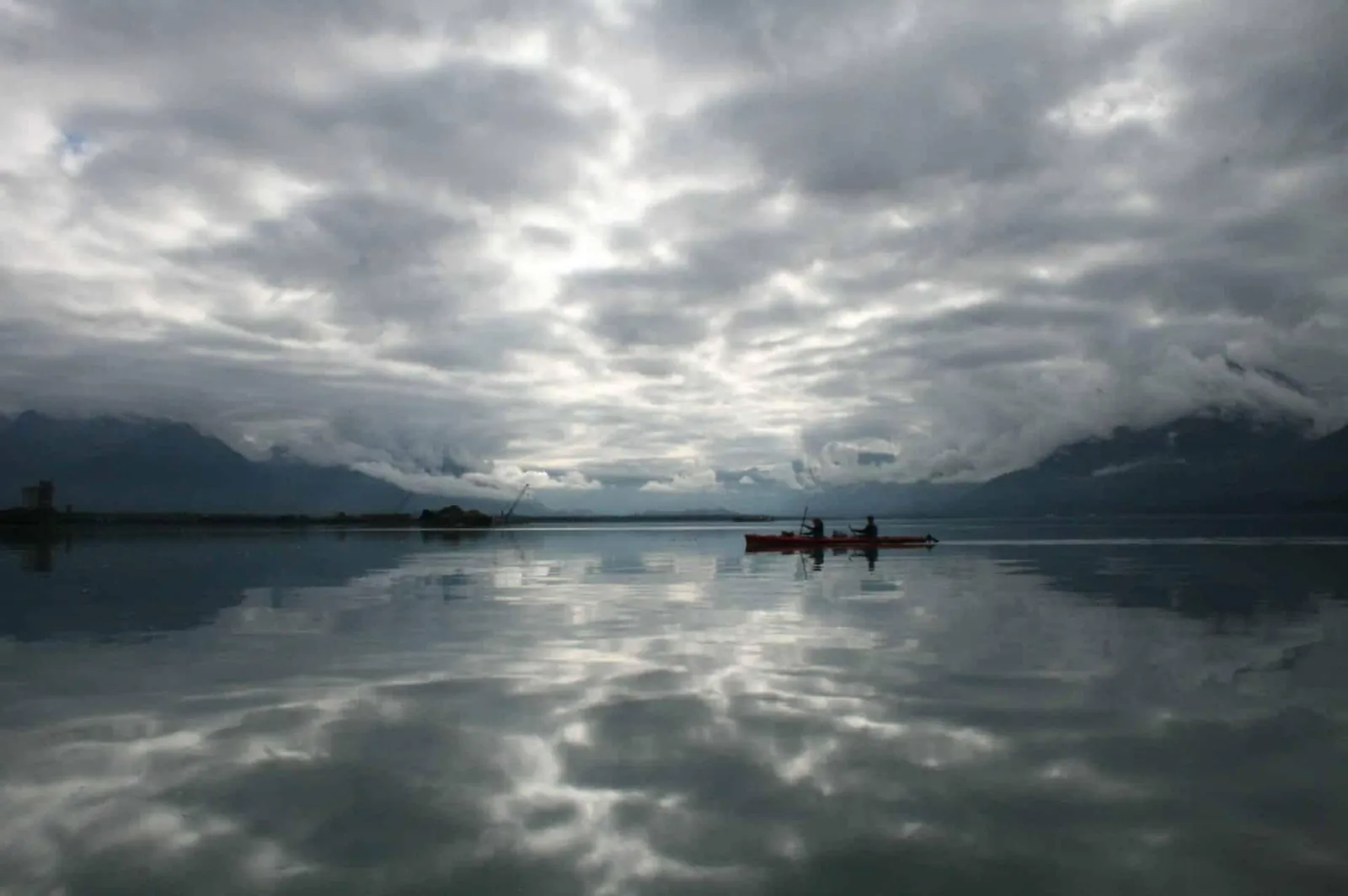 A single kayak on mirror-flat water under heavy cloud in Port Valdez